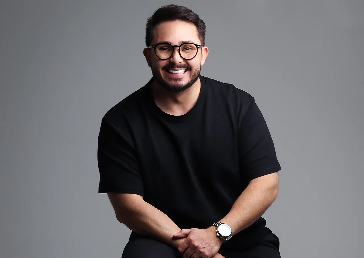 Kleber Toda sentado, sorrindo, com camiseta preta e óculos, diante de fundo cinza
